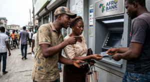 A photograph showing an elderly woman in traditional clothing at an outdoor ATM. She is being approached by a man in a cap, who is aggressively pointing at the ATM screen, and a second man who is attempting to hand her a plastic card while she looks confused. The woman appears confused and distracted at an ATM as two men, potentially scammers, simultaneously point at the screen and attempt to hand her a card.